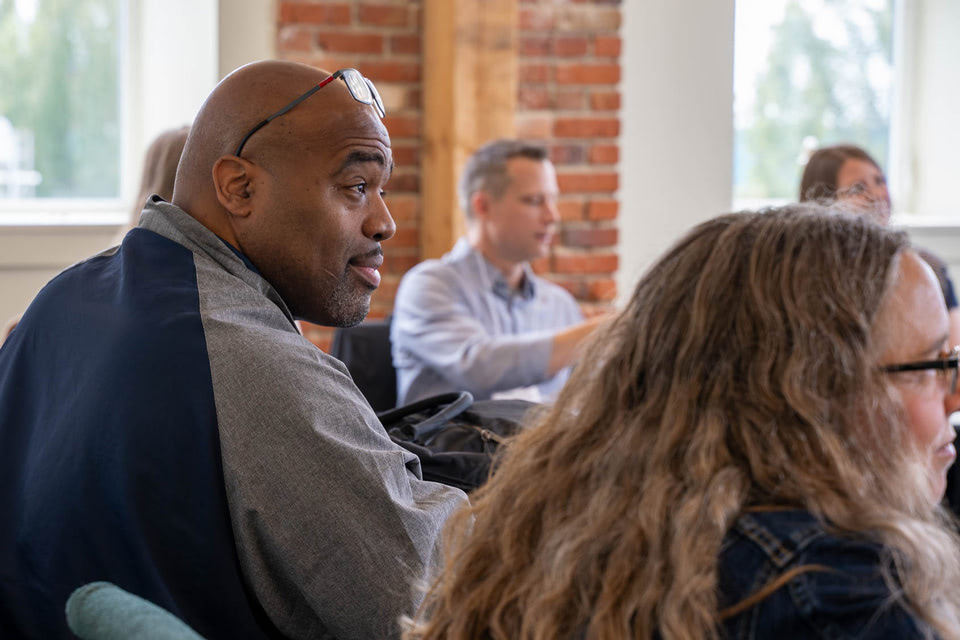 People sitting in a meeting or workshop, with one man in focus wearing glasses on his head and listening attentively learning about Narrative Focused Trauma Care®