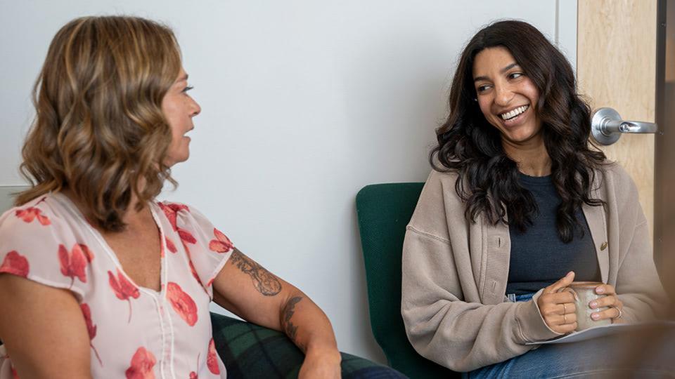Engage Your Story, Two women sit and talk warmly together in a casual setting. One woman smiles while holding a mug and a notebook, and the other listens attentively